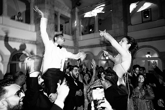 Wedding dance party with guests raising hands and cheering, some filming on smartphones, in a grand ballroom with arched doors and balcony