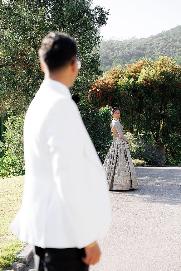 First look moment as groom seeing bride in a lehenga walking away on a paved garden path with flowering bushes and hillside behind