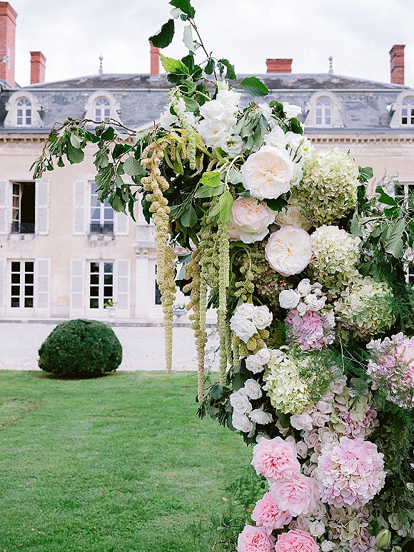 Wedding floral arch with hydrangeas and roses, asymmetrical greenery and hanging amaranthus on a lawn before a manor house facade