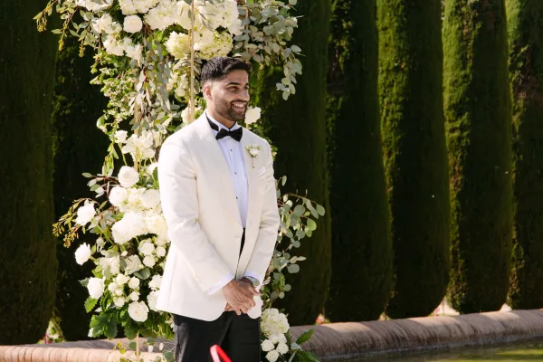 Groom portrait in a white tuxedo groom look with black bow tie and rose boutonniere, standing beneath a floral arch in a garden setting