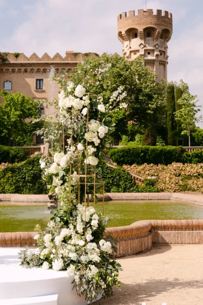 Ceremony floral arch of white flowers and greenery on a gold metal frame by a fountain, with a castle garden and pond behind
