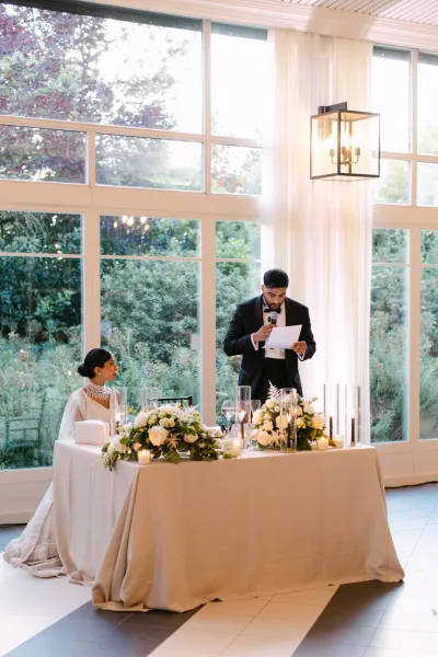 Wedding reception toast as the groom in a tuxedo speaks into a microphone while the bride listens at a candlelit head table by large windows