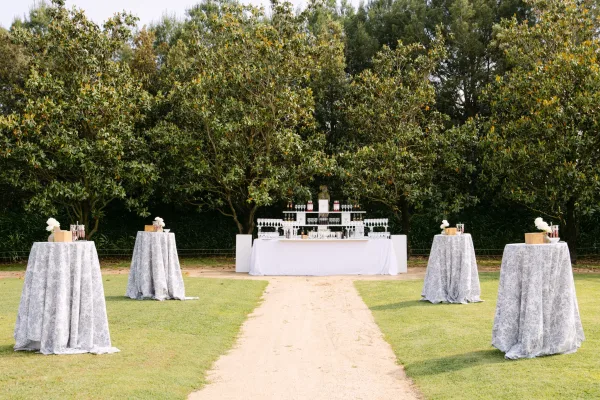 Wedding bar setup with a white bar front, tiered bottle display, and glassware on a garden lawn beside cocktail tables with floral accents