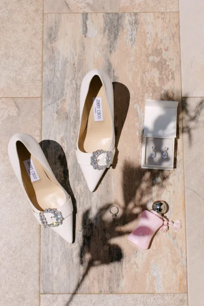 Bridal accessories flatlay with white wedding heels and crystal buckles, drop earrings, ring, and pink perfume on stone tiles in light shadows