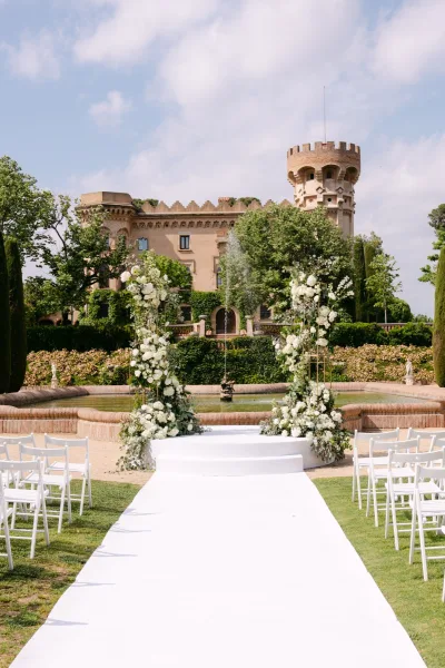 Ceremony setup for an outdoor wedding ceremony with white aisle runner leading to round platform, white floral pillars, and fountain at historic villa garden
