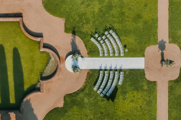 Outdoor ceremony setup with a white aisle runner leading to pedestal florals, curved chairs on a lawn by a pond and stone paths