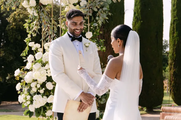 Wedding vows as bride reads from vow cards into a microphone under a white rose floral arch, groom in white tux reacts in sunlit garden