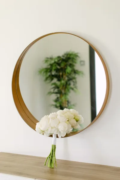 Bridal bouquet of white peony blooms with ribbon-wrapped handle resting beside a brass-framed round mirror on a wooden tabletop