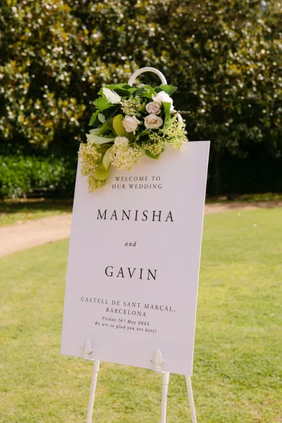 Wedding welcome sign on a white easel with white rose floral arrangement, set along a garden pathway with trees and lawn grass