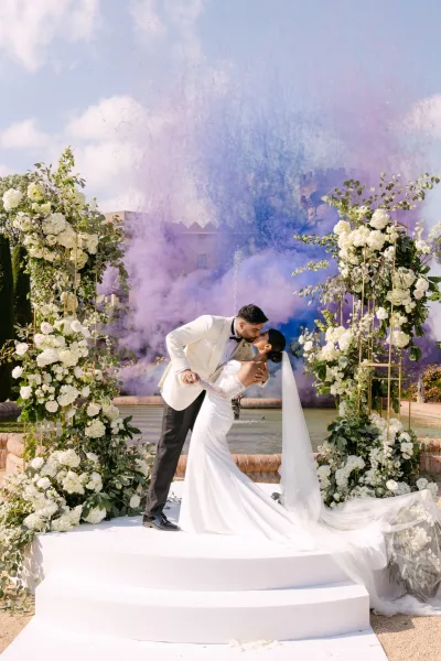 Wedding kiss as groom dips bride in strapless gown and long veil under a white rose floral arch, with fountain and colored smoke behind