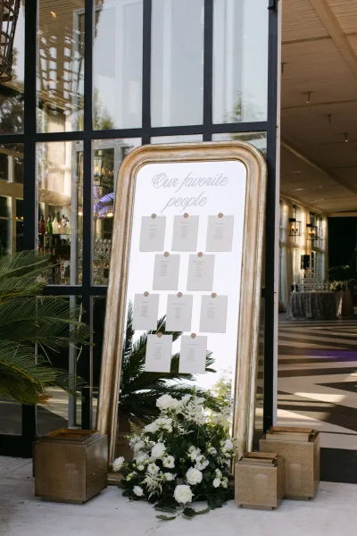 Wedding seating chart in an arched gold frame with wax-sealed cards, white florals and lanterns by black-framed glass windows indoors