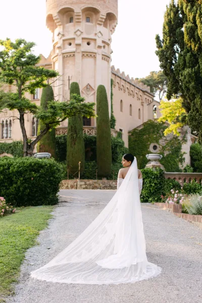 Bridal portrait of a long veil bride looking over her shoulder, lace sleeves and earrings, on a gravel path by a castle garden hedge