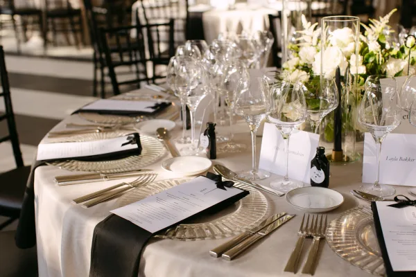 Reception tablescape with wedding table setting of white roses and greenery in glass vases, taper candles, and black ribbon menu cards on round tables