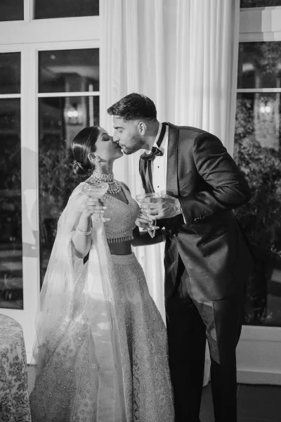 Wedding kiss portrait of newlyweds kissing while holding champagne coupes, bride in veil and jewelry beside tuxedoed groom near tall windows