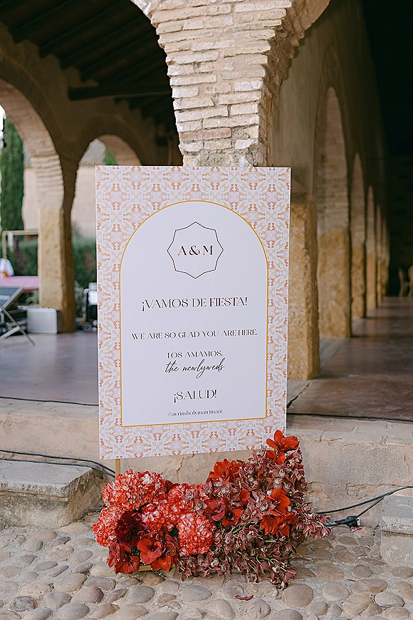 Wedding welcome sign on a stand with red floral arrangement accent, set under stone arches along a cobblestone walkway