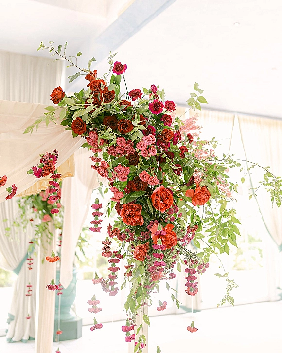 Wedding floral arch with ceremony arch flowers, roses, hanging blooms, and greenery with draped fabric in a bright indoor venue by windows