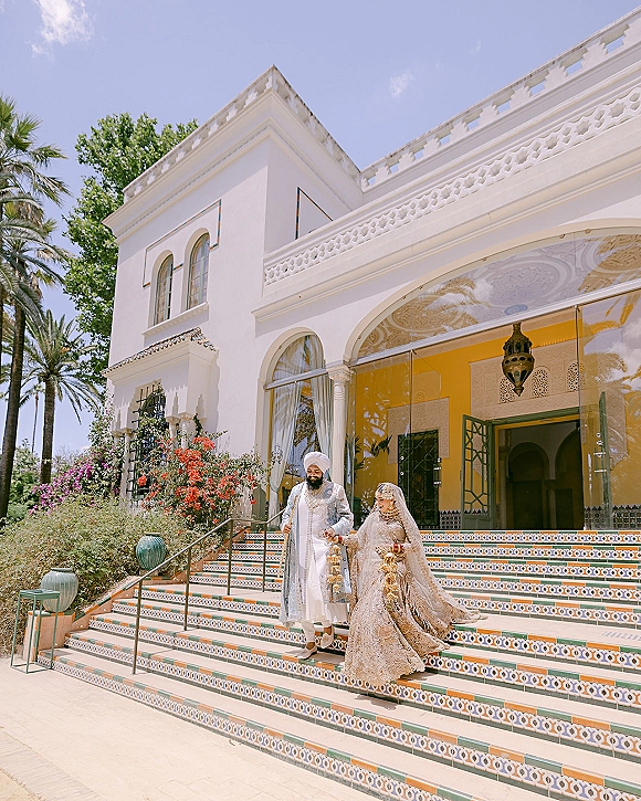 Wedding couple portrait of a bride and groom walking down stairs, holding hands, with bridal veil against white stucco arches and palm trees