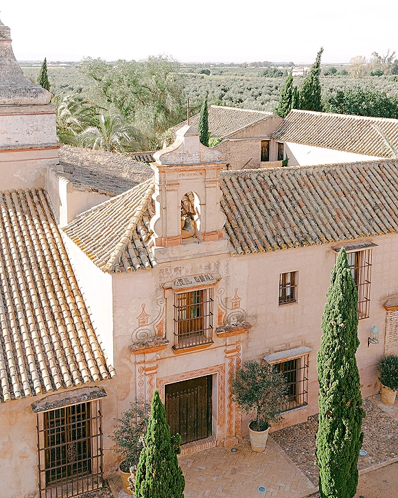 Wedding venue exterior with a clay tile roof and bell tower, stucco facade and arched doorway, framed by cypress trees and olive groves