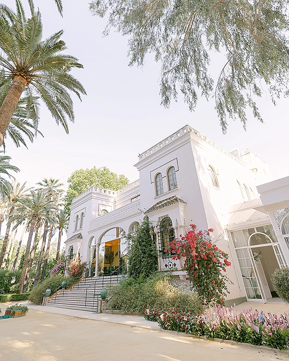Wedding venue exterior with arched windows and a grand staircase with wrought iron railings, framed by climbing vines and palm trees