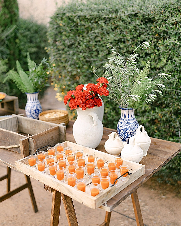 Welcome drinks station with wedding welcome drinks in shot glasses on wooden trays, styled with ceramic pitchers and red flowers by garden hedges