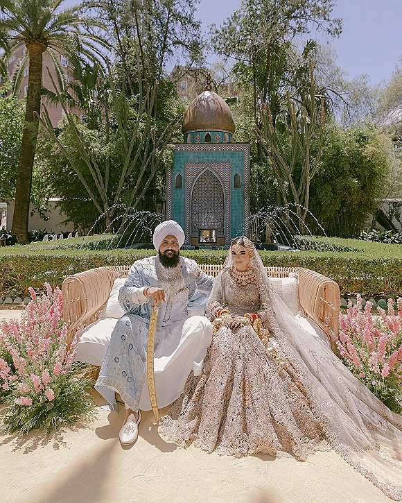 Wedding couple portrait of a South Asian bride and groom seated on a wicker sofa, her silver lehenga and his turban before a garden fountain backdrop