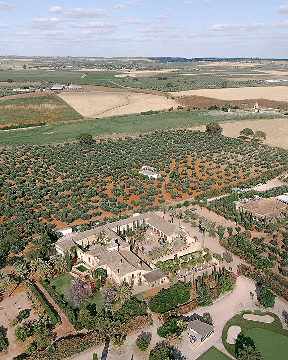 Wedding venue aerial view of a villa estate with palm-lined courtyard, gardens, and gravel driveway amid orchards and rolling hills