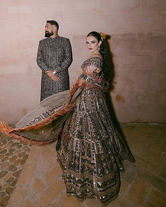 Couple portrait of a South Asian bride in a black and gold lehenga and groom in embroidered sherwani against a stone wall backdrop