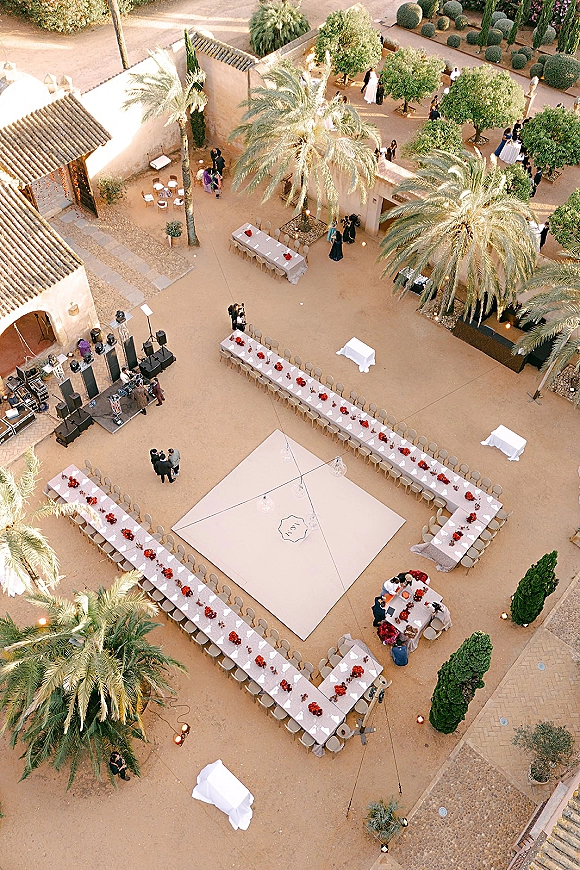Outdoor reception setup with U-shaped long banquet tables in white linens, red floral centerpieces, dance floor and stage in a palm-lined courtyard