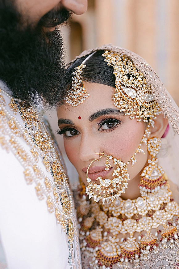 Bridal portrait of a south asian bride portrait with nath nose ring chain and gold jewelry, leaning on groom against a neutral backdrop