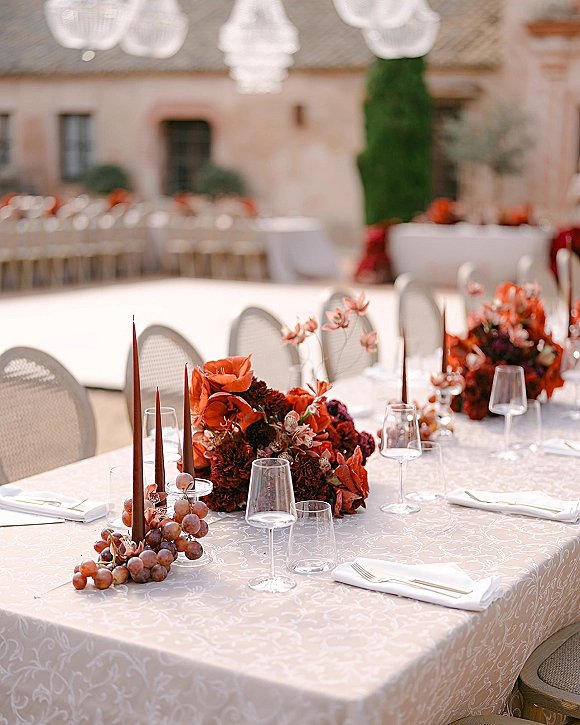 Reception tablescape with an outdoor reception table set with red floral centerpieces, taper candles, grapes, and glassware in a courtyard
