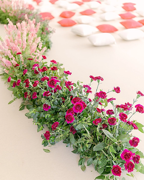 Ceremony aisle decor with ground floral aisle meadow of pink and magenta blooms, greenery garland, and white and red floor pillows on a light runner indoors