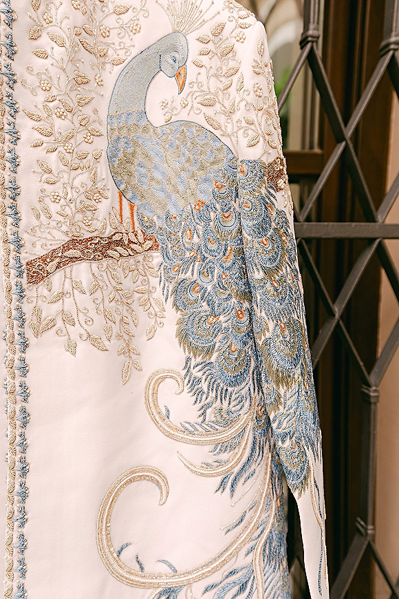 Embroidered wedding outfit with peacock embroidery design and intricate beadwork, shown hanging beside a window frame indoors