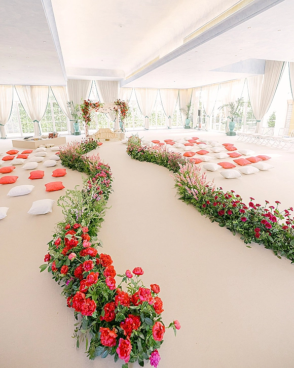Ceremony aisle decor with a floral lined aisle, ground flower arrangements and floor cushions beneath a floral arch in a bright windowed hall