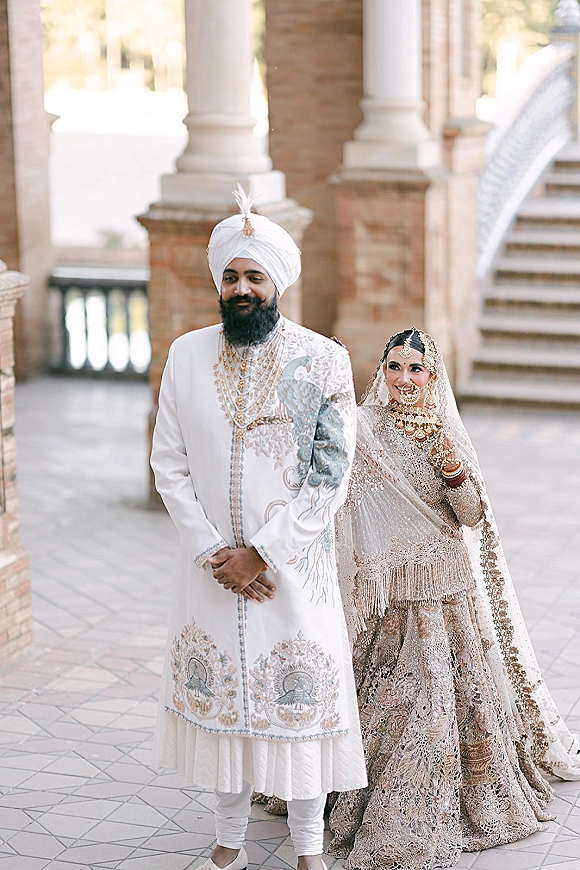 Couple portrait of a South Asian wedding couple in embroidered lehenga and sherwani with bridal veil, under a stone archway by brick columns