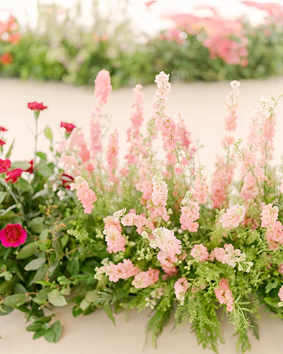 Wedding floral arrangement with pink snapdragons and red roses, lush greenery and ferns, set against blurred garden foliage and a neutral wall