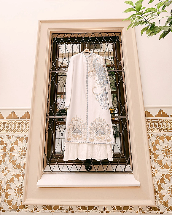 Groom wedding outfit, an embroidered sherwani on a wooden hanger by a window grille, gold and blue embroidery against cream walls and tile
