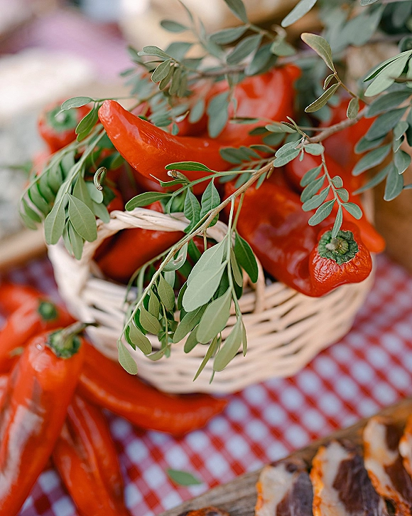 Wedding food display of red peppers and greenery sprigs in a wicker basket on a gingham tablecloth beside a wood serving board