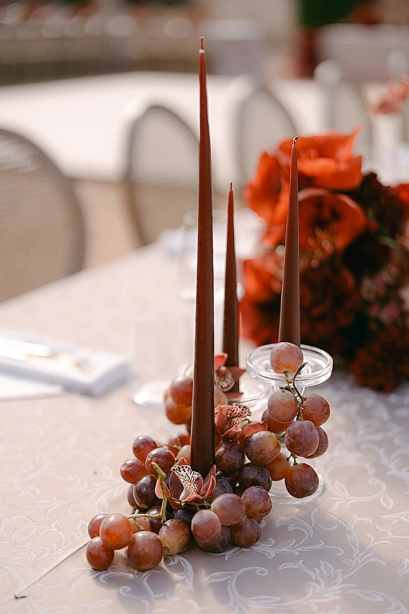 Reception tablescape with wedding taper candles, grape clusters, and a floral centerpiece on a patterned tablecloth in outdoor light