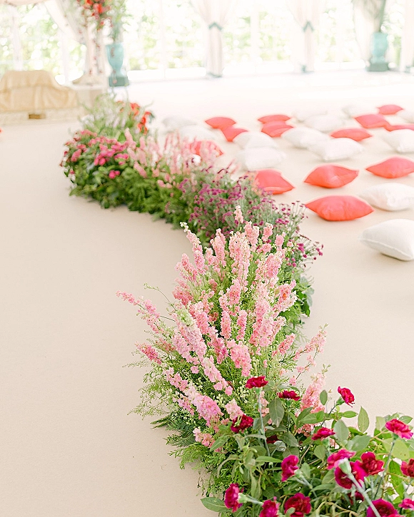 Ceremony aisle florals border the white floor with pink and red blooms, greenery and floor pillows under light tent draping by bright windows