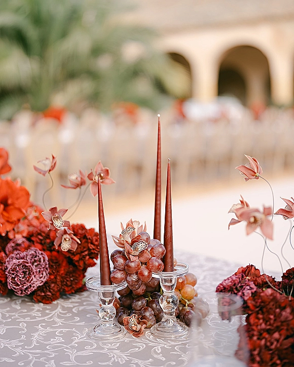 Wedding tablescape with a red wedding centerpiece of orchids and grapes, plus taper candles in glass holders on patterned linen outdoors