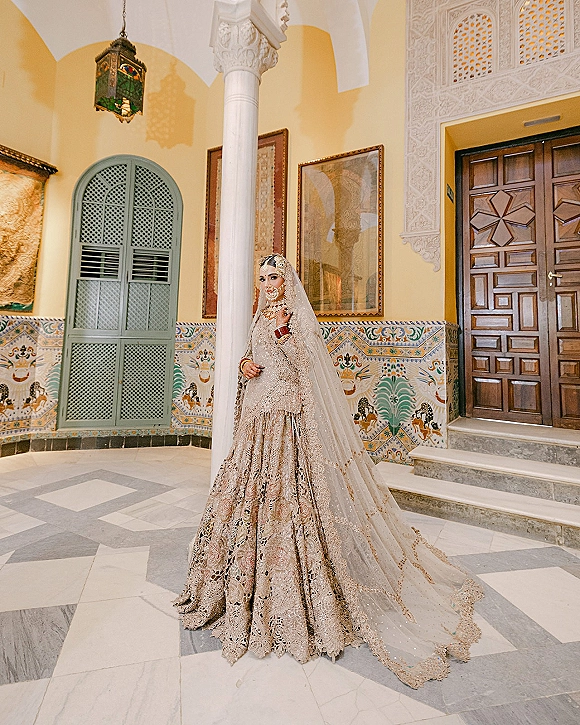 Bridal portrait of a bride in an embellished bridal lehenga look with a sheer veil, ornate jewelry, standing by an arched doorway