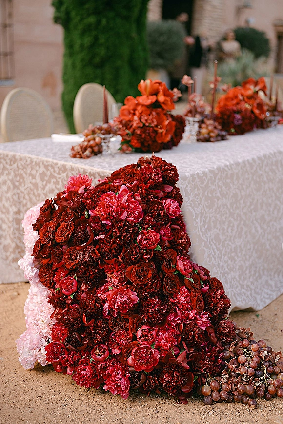 Reception tablescape with wedding head table decor, lace tablecloth, burgundy florals, taper candles, grapes and glassware in a stone courtyard