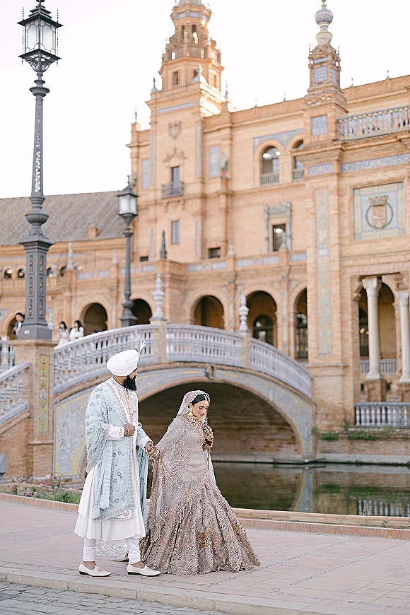 Couple portrait of a south asian wedding couple holding hands, bride in embellished lehenga and veil beside a canal and arched bridge backdrop