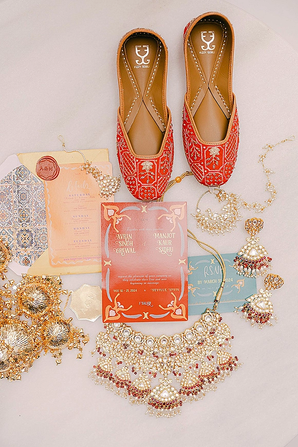Wedding flat lay featuring red embroidered jutti shoes, invitation suite with wax seal, and pearl and gold jewelry on a neutral surface
