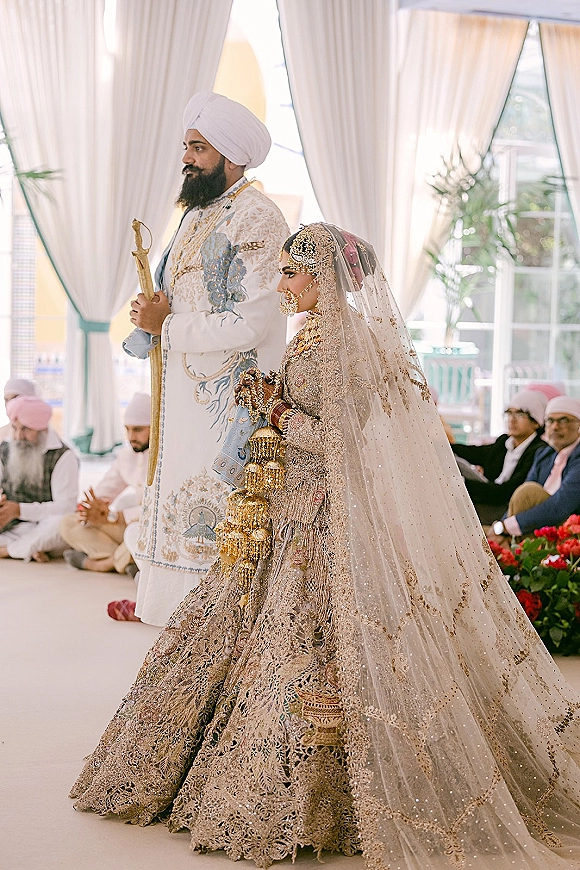 Ceremony moment at a Sikh wedding ceremony with bride in lehenga and groom in turban walking past seated guests under white drapery