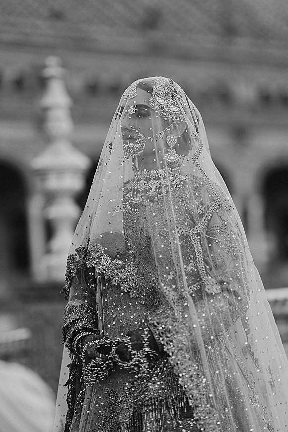 Bridal portrait of a bride wearing veil over her face, showcasing a beaded veil, jewelry and gloves on an outdoor terrace by a stone railing