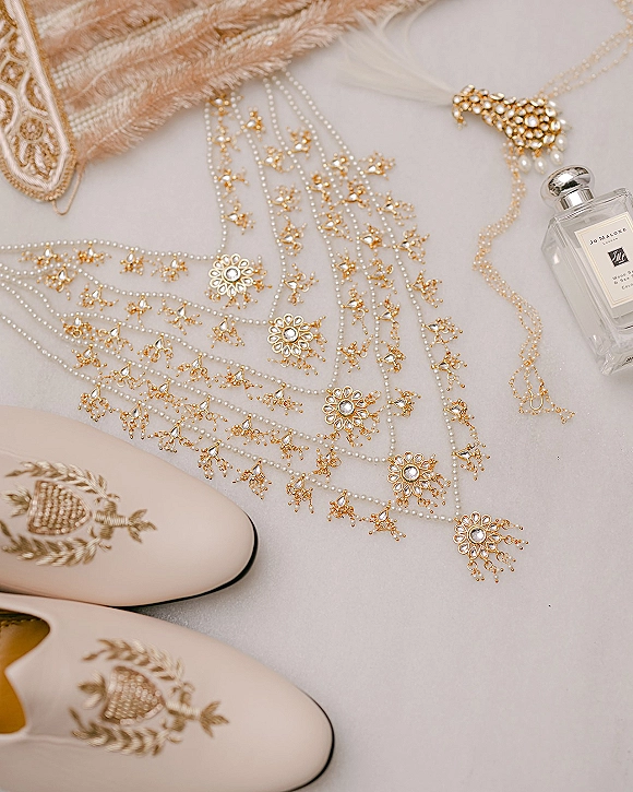 Bridal jewelry flatlay with pearl bridal jewelry, gold floral pieces, embroidered shoes and perfume on light fabric with pampas grass accent