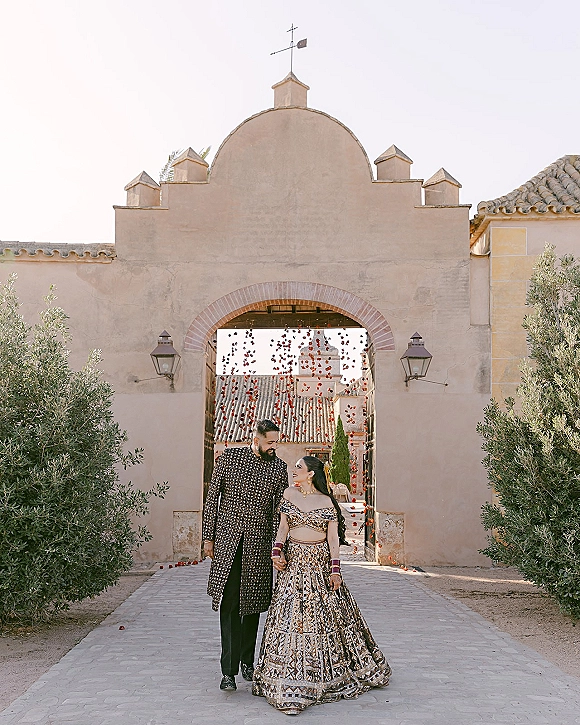 Couple portrait of an Indian wedding couple holding hands, bride in gold lehenga and groom in black sherwani under a stucco archway courtyard
