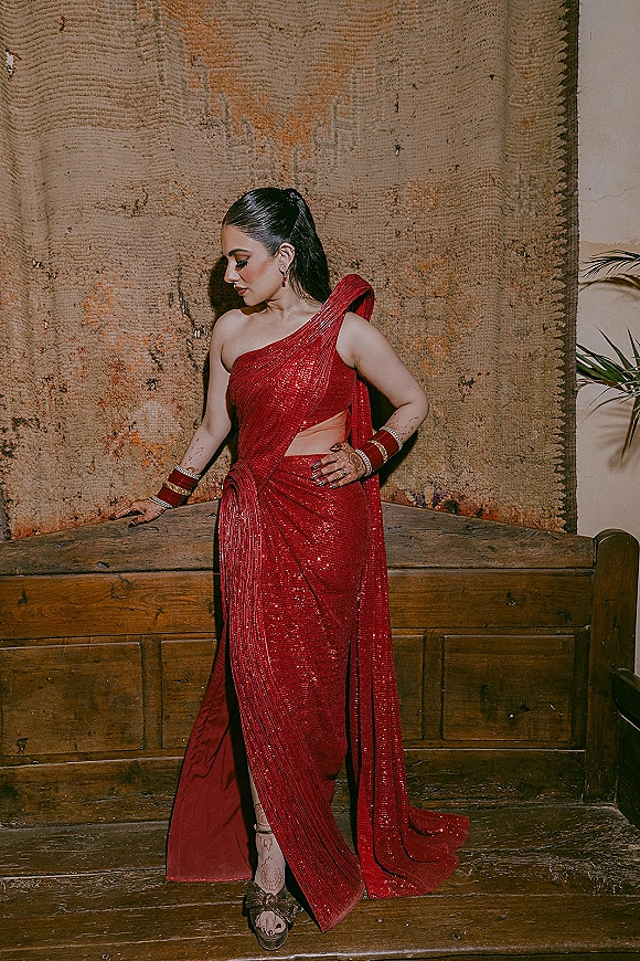 Bridal portrait in a red sequin saree, shown in side profile with hennaed hands and bangles beside a wooden bench indoors