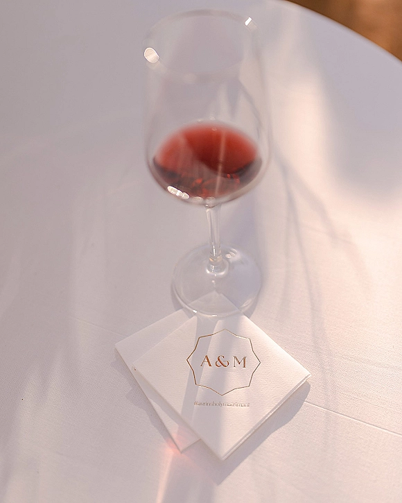 Wedding cocktail napkin with custom monogram napkins in gold foil beside a wine glass of red wine on a sunlit white tablecloth